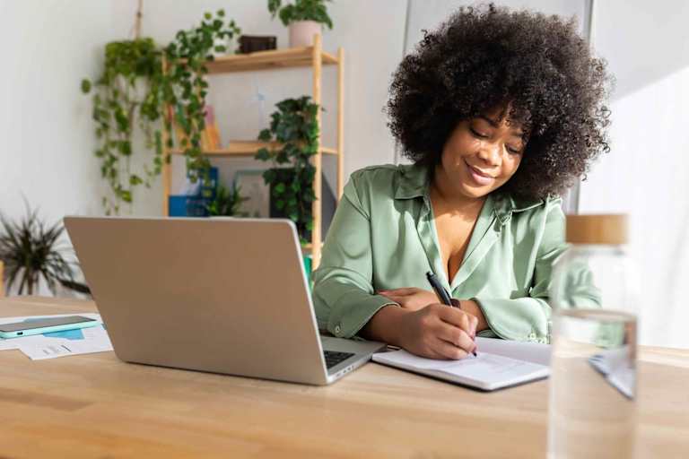 Psychology graduate student writing on notebook while using laptop computer.