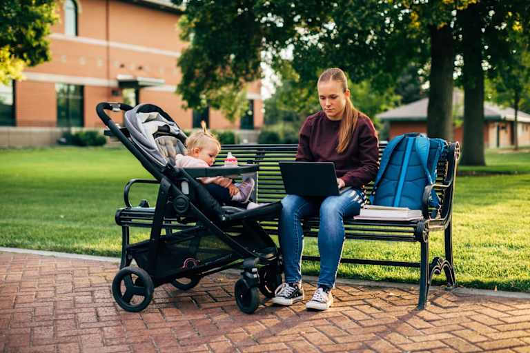 A mother sits on a park bench with her baby daughter in a stroller next to her. She is on a university campus working on her laptop.