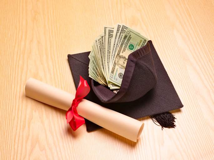 A rolled up diploma laying on a table next to a graduation cap filled with U.S. cash.