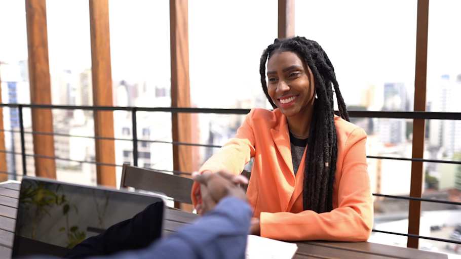A Black female college student sitting down in a corporate office shakes hands with a male interviewer off screen.