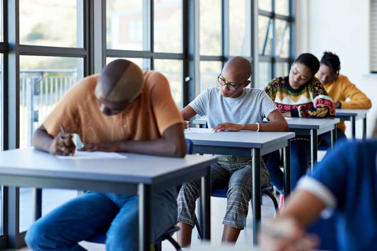 Young male and female university students writing on papers at desks by window in examination hall