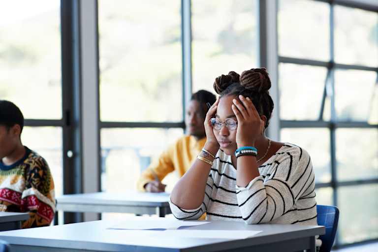 Worried young female student sitting with head in hands at desk during exam in community college classroom