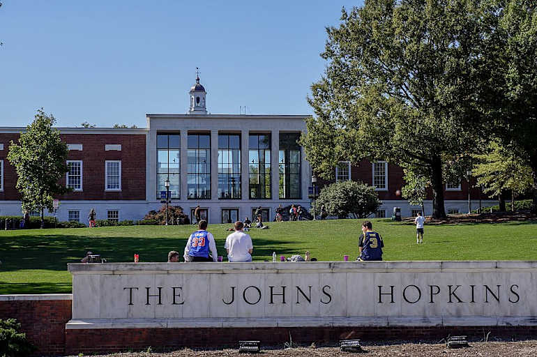 A view from Charles street of the grassy "Beach" and Milton S Eisenhower Library of the Johns Hopkins University; three male students, their backs facing the camera, sit on top of the marble university sign, other students sunbathing and playing catch on the grassy hill; the clock tower of Gilman Hall, a humanities building, peeks above the top of the library; Baltimore, Maryland, March, 2014. Courtesy Eric Chen. (Photo by JHU Sheridan Libraries/Gado/Getty Images).
