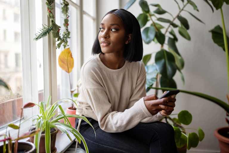 Woman sitting on the windowsill with mobile phone and looking away. Woman relaxing at home looking outside the window.