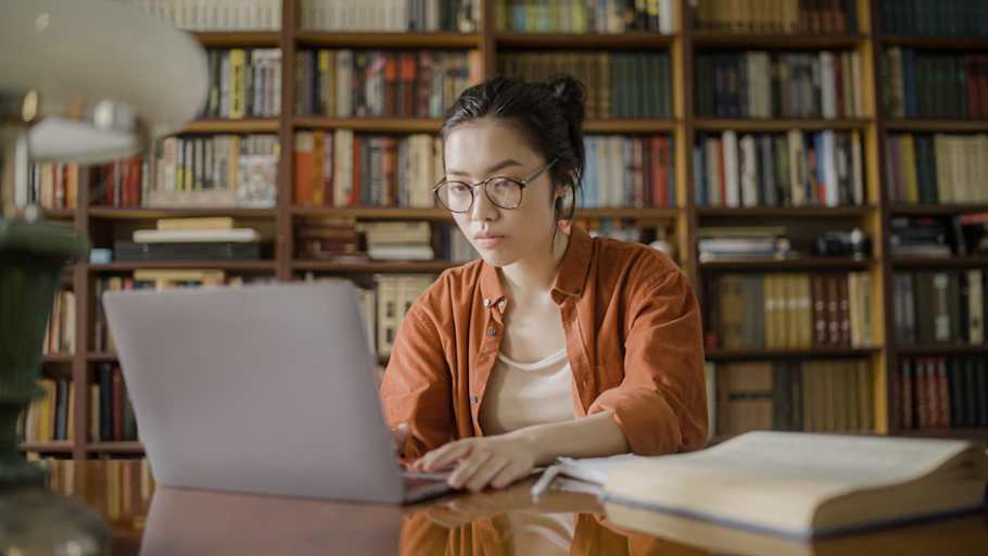 Focused Asian American female college student doing research in the library, typing an article on her laptop.