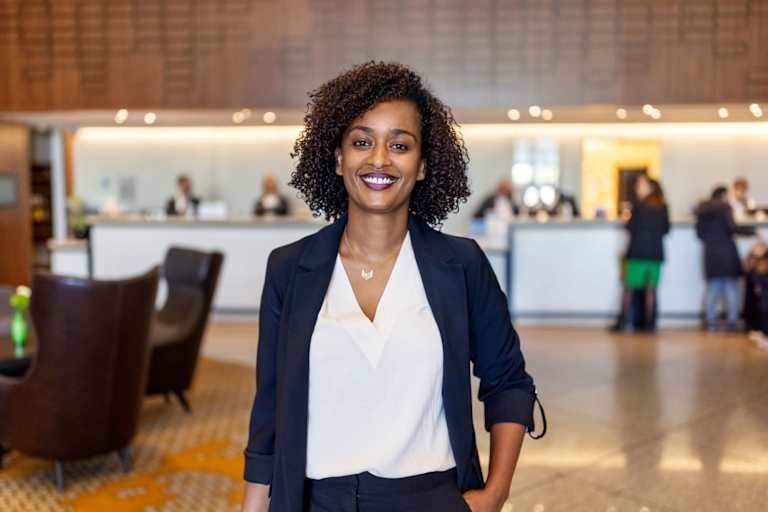 Black female hotel manager standing in the middle of a hotel lobby. She is looking towards the camera and smiling.