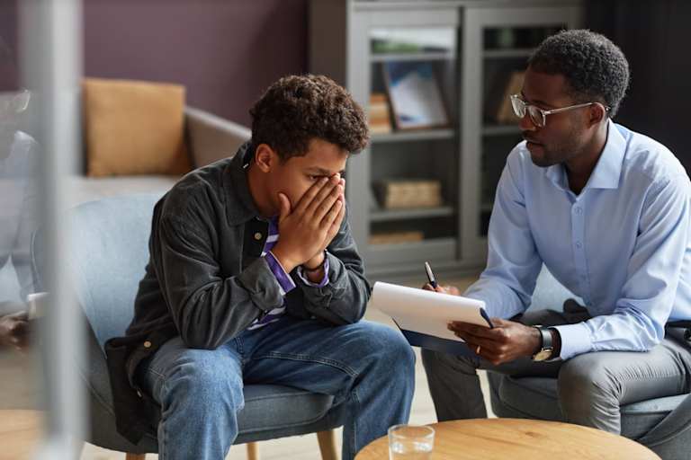 Black teenage boy crying when sharing his experiences with a Black male adolescent psychologist.
