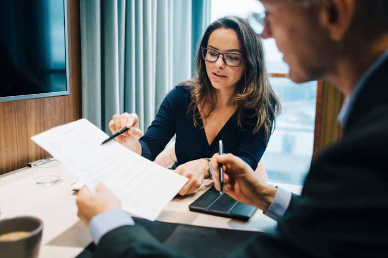 A Caucasian female business lawyer is explaining details on a paper contract with a male client in her office.