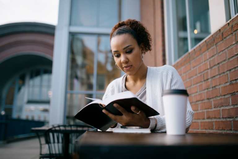 A mixed race female college student reading a textbook while sitting outdoors at a cafe on campus.