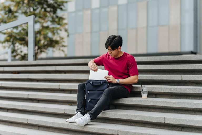 Male Asian American college student sits on stairs outside of a university campus building during his break. He is packing away his laptop in his backpack before heading off to his next class.