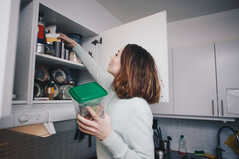 Female college student searching for ingredients in her apartment kitchen shelves.