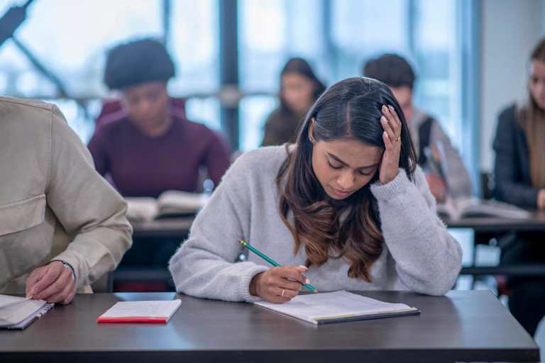 A female college student sits at her desk in a university classroom and takes notes. She has her head in her hand in concentration.