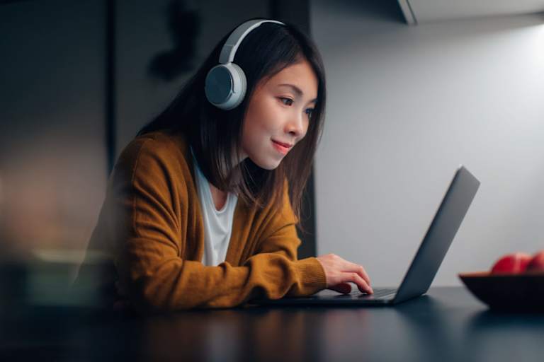 Asian American female college student wearing headphones, studying online on her laptop at home during the night.