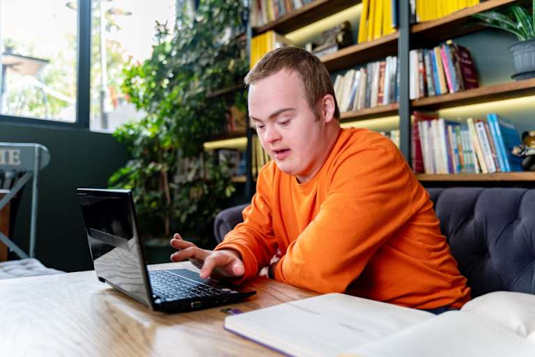 Male college student with Down syndrome sitting at a table in his living room. He is typing on his laptop while studying for a class.