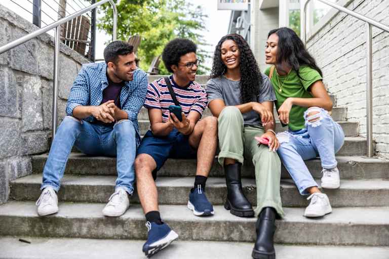 Group of college friends sitting on steps look together at a smartphone and smile.
