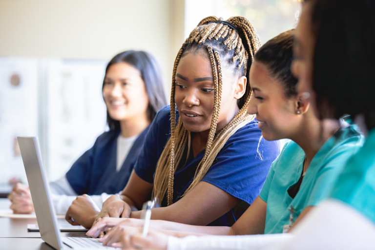 A diverse group of four female nursing students sitting in a college classroom. They are discussing coursework with each other. One of the students is writing notes on her laptop.