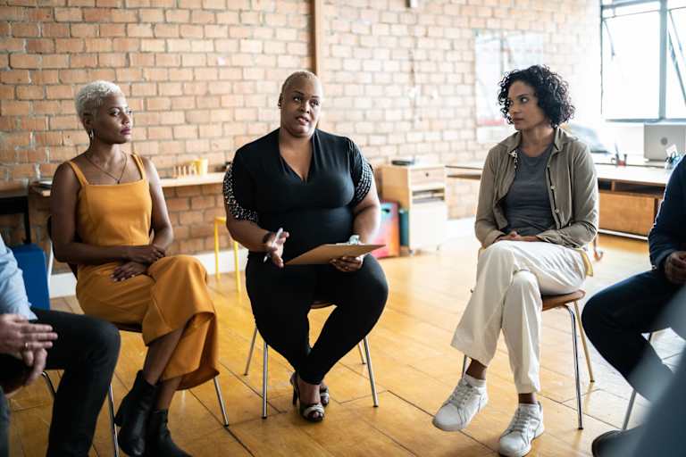 A Black mid-adult female social worker leading a group therapy session for people of color in the community. She is sitting at the head of the circle for the group and holding a clipboard for notes.