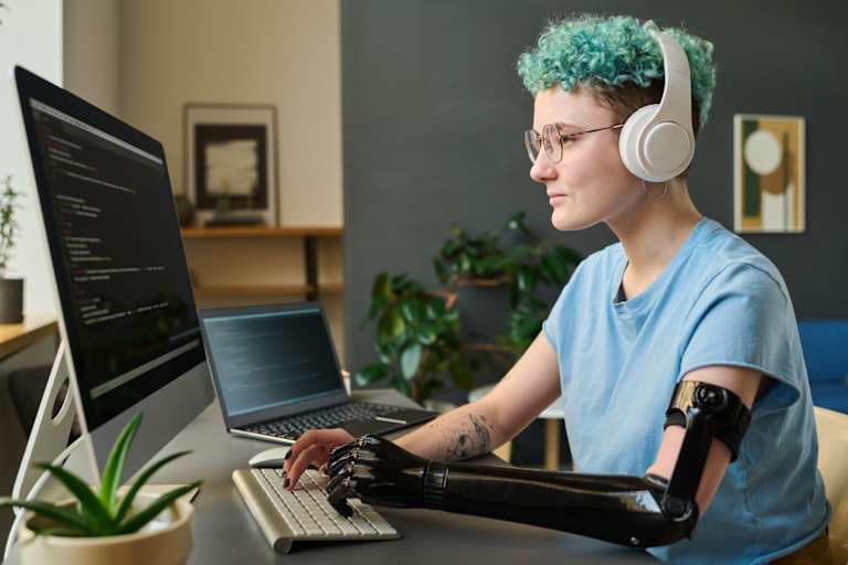 Female computer programming student with a prosthetic left arm sitting at her living room desk at home. She is wearing wireless headphones and testing a security program on her desktop computer for a class assignment.