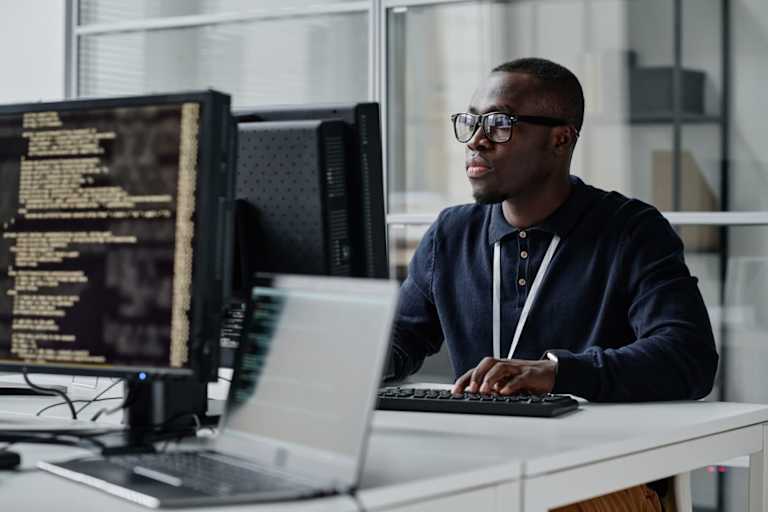 Black male cybersecurity engineer sitting at his desk in a corporate office. He is typing code for an internal security program on his desktop computer.