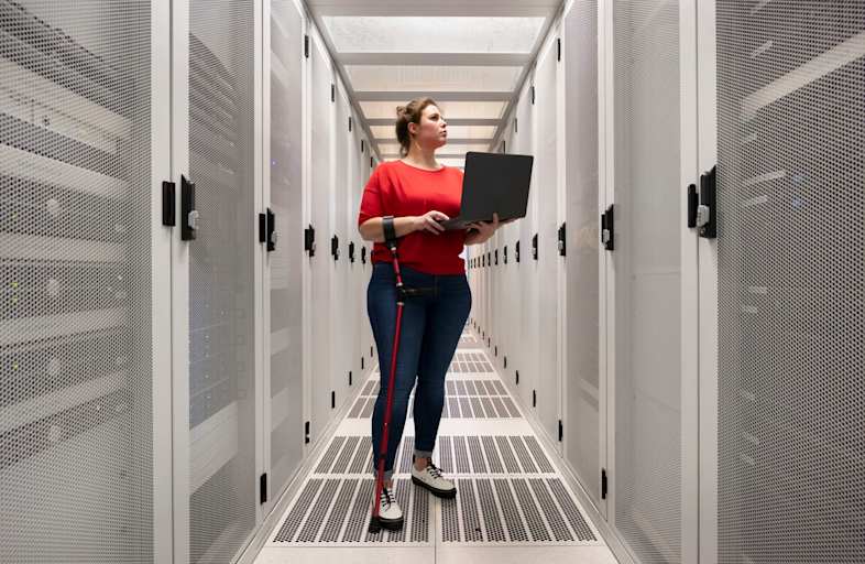 Female cybersecurity engineer with a forearm cane standing in a corporate data center. She is examining the computers while referencing information on her laptop.