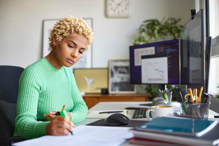 Black female business professional working from home at her desk. She is writing notes with a felt tip pen on paper while looking at charts and spreadsheets on her desktop computer.