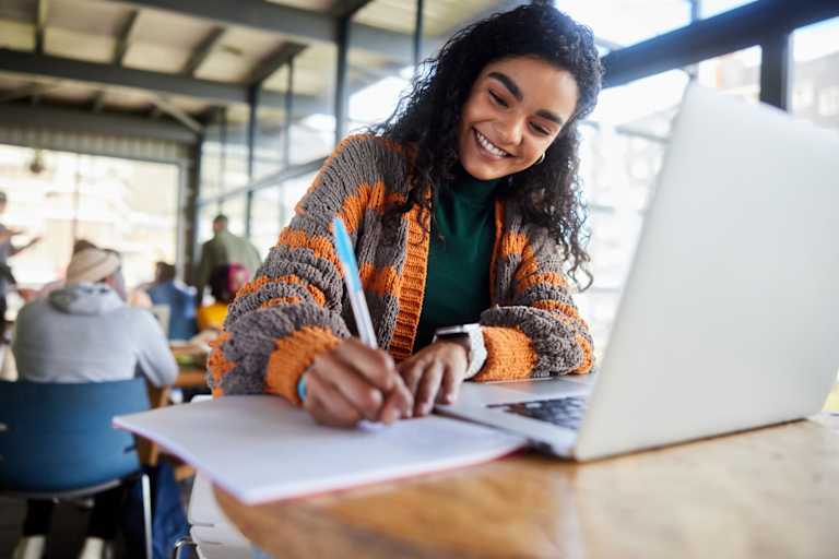 Smiling Black female college student writing notes and working on a laptop while doing her homework in a busy campus cafeteria.