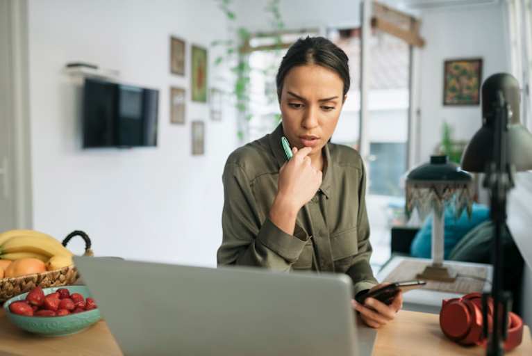 Woman sitting at home looking at her calendar on her laptop. She is holding her smart phone in her left hand, trying to reschedule her job interview.
