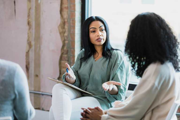 A mid adult Black female psychologist gestures as she speaks to a young adult woman during a group therapy session for women of color.