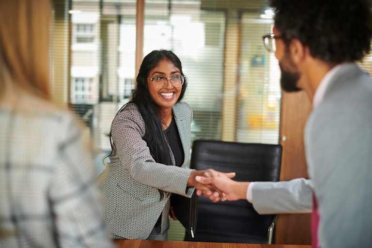 Female South Asian college graduate shakes hands with two employees during an onsite interview at a corporate office.