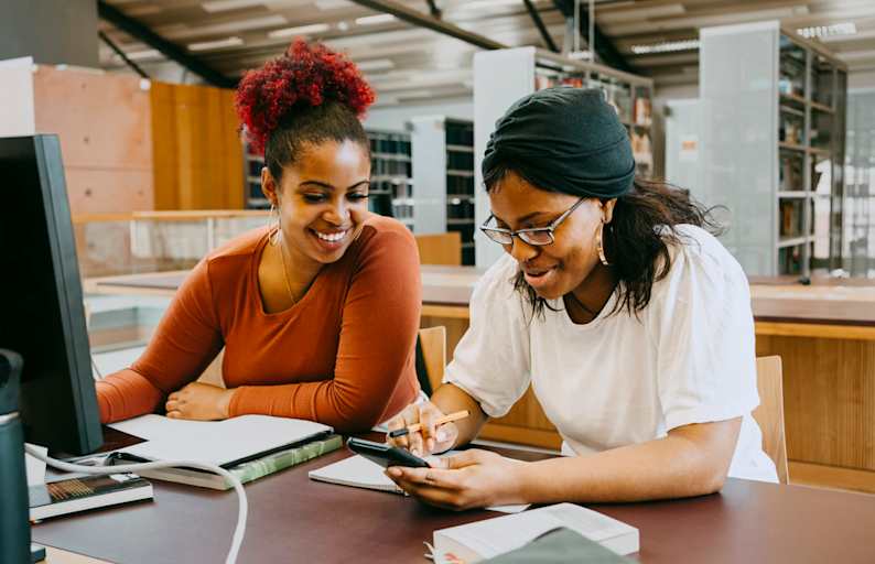 Two smiling Black female students sitting next to each other at a library desk on campus. They are researching information using one of the library's desktop computers during a study session.
