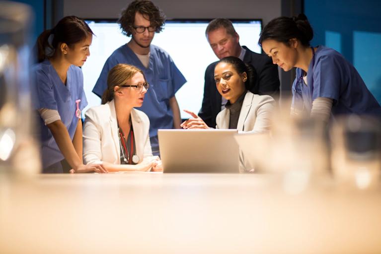 Black female healthcare administrator leads a business meeting with doctors and nurses in a hospital meeting room. She is showing them data on her laptop.