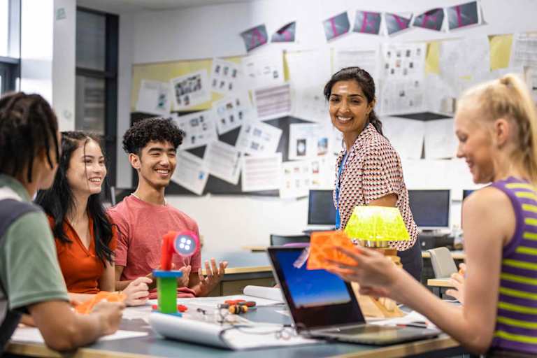 A female South Asian American high school teacher sitting with her students in class, discussing their work. They are all wearing casual clothing and smiling.