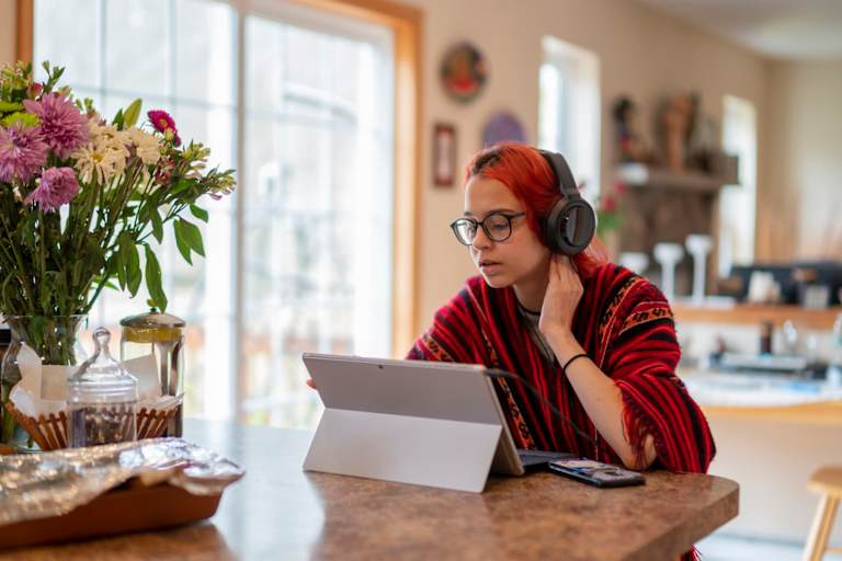 Teenage girl wearing poncho and headphones is taking a distant class, working with a laptop computer at the kitchen counter.