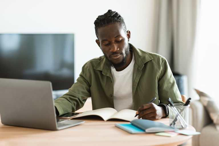 Focused african american man sitting at desk working on laptop and taking notes in notebook, studying online at home