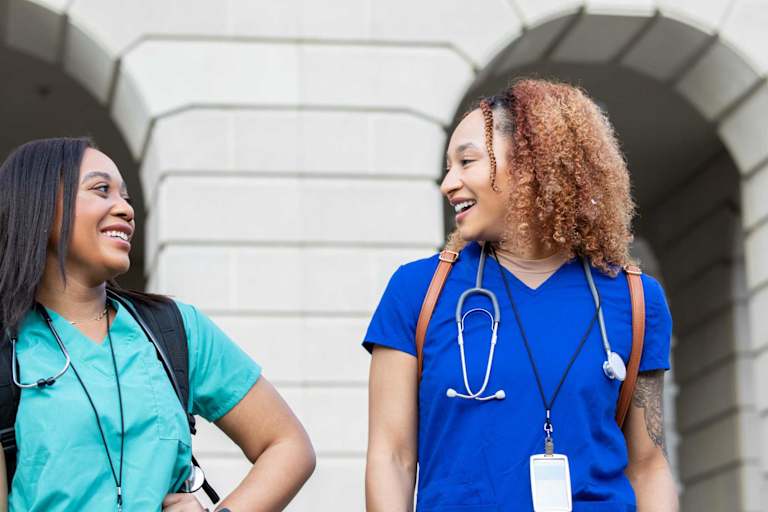 Two Black female medical school students laugh while walking on campus. They are wearing blue medical scrubs and carrying backpacks.