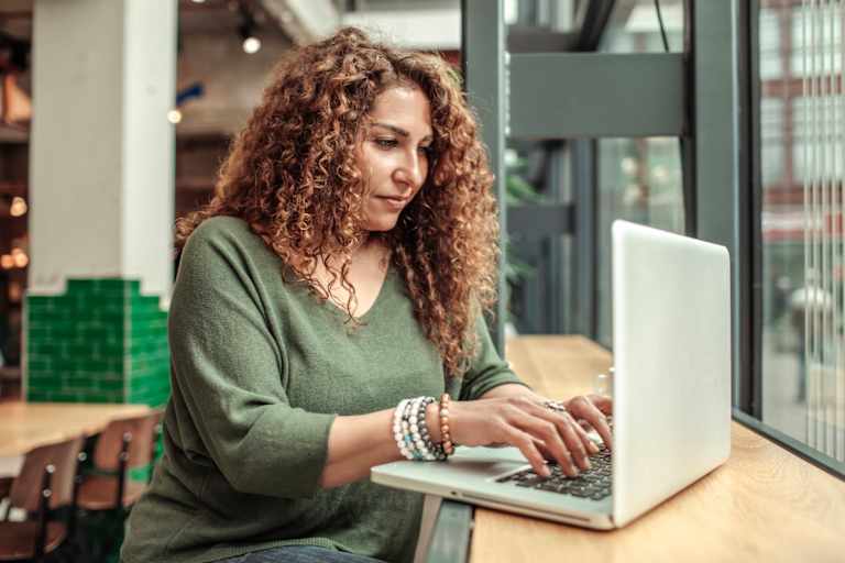 Woman in her mid-40s sitting in a cafe. She is typing on her laptop, as part of an assignment for an online course.
