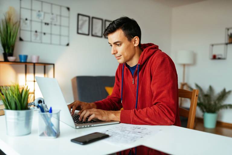 Male college student sitting at a desk at home in his living room. He is typing a report on his laptop for a class assignment.