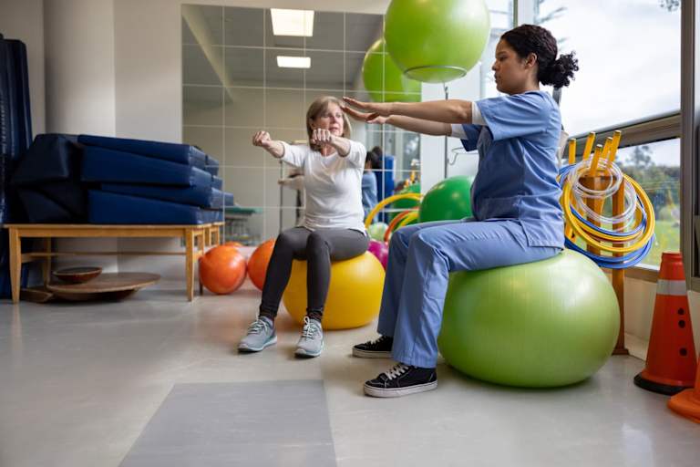 A Latina physical therapist meeting with a senior female client during a PT session at a sports clinic office. They are sitting on green plastic stability balls. The therapist is leading her client through some arm exercises to do at home.