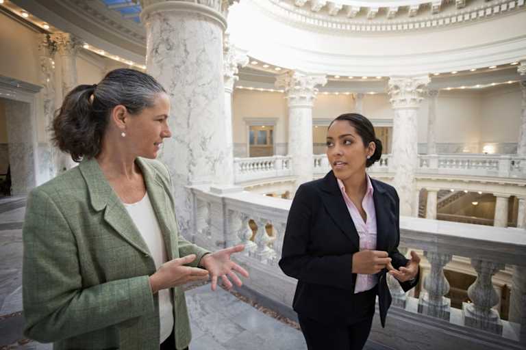 Two female politicians, one a Caucasian woman in her late 40s and the other a younger Black female, are talking while on their way to a meeting in a government building.
