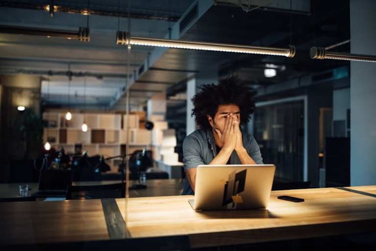 A young Black male employee sitting at his desk in a corporate office late at night. He has his hands on his face and his laptop is open.