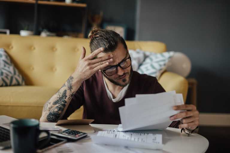 Caucasian male college student sitting at his living room coffee table. He has a worried look on his face while he's holding several paper bills in his hand.