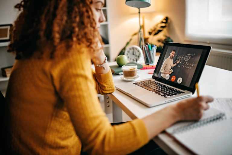 Caucasian woman sitting at a desk in her apartment. Her laptop is open, and she is taking notes on a notepad. She is taking a class online: her professor appears in a Zoom meeting on her laptop, and is demonstrating math concepts on a blackboard behind him.