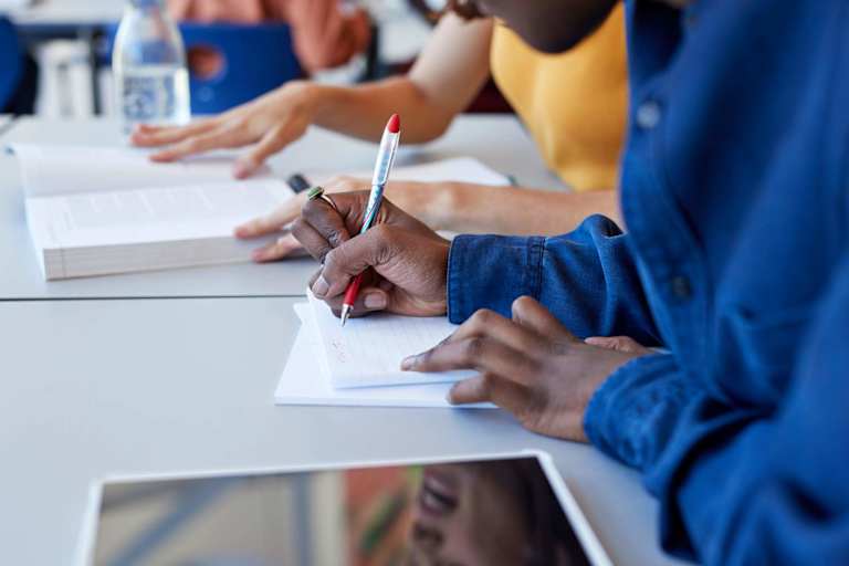 Close up of a Black male high school student writing in a notebook during a study session with a female friend. They are sitting at a table in a high school lunch room and looking at class textbook materials.
