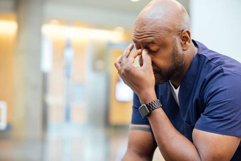 Tired Black male nurse sitting in a chair in the hospital break room. He is rubbing his temples.