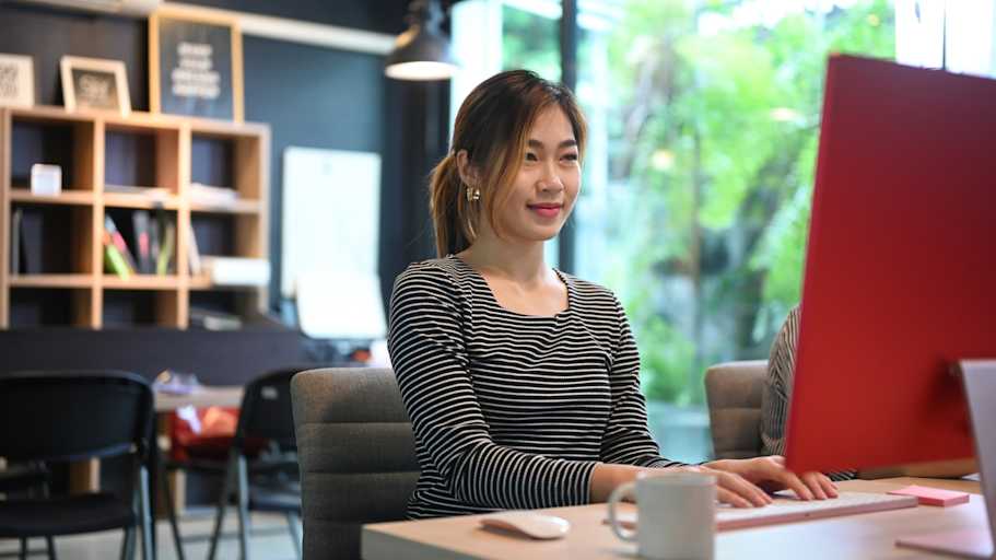 Asian American female student sitting at a desk in a coding bootcamp classroom. She smiles as she types a software program on her desktop computer.