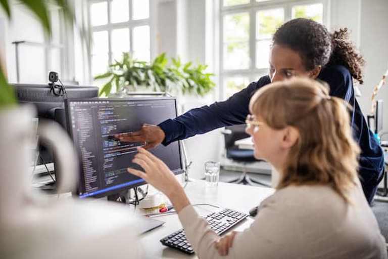 Lady pointing to a computer monitor showing her coworker how to troubleshoot code