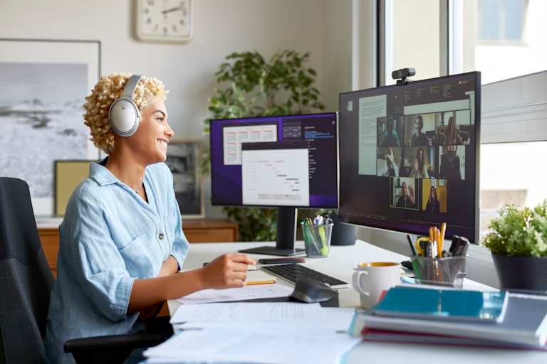 Smiling Black female student chatting with her classmates over video before an online class. She is sitting at a desk in her home office wearing wireless headphones.