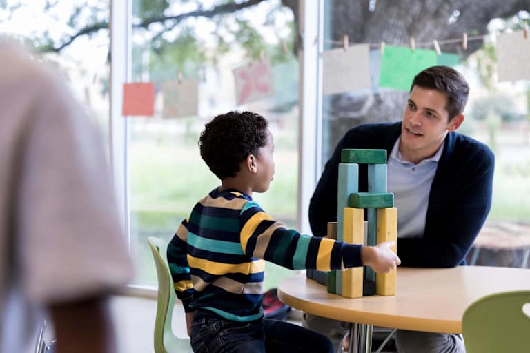 The young boy and the mid adult male teacher sit together at a table and talk.