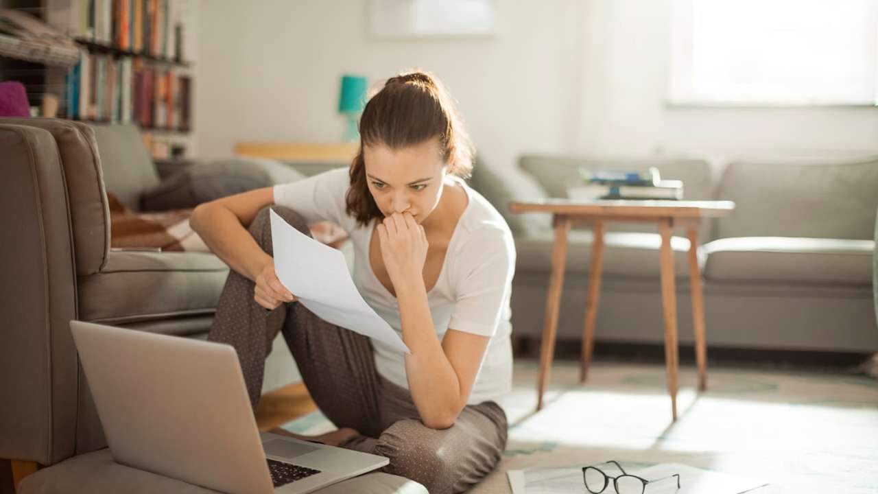 Photo of an anxious college student sitting on the floor in her living room. She has her laptop open and a college tuition bill in her hand.