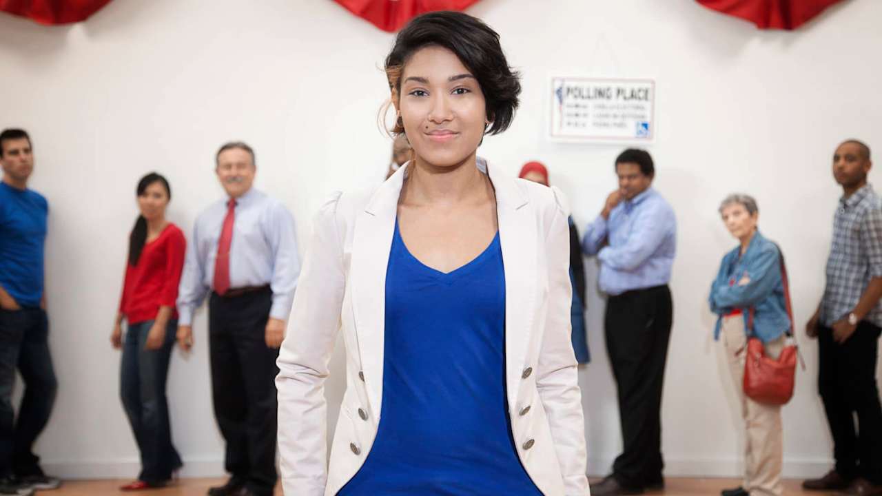 Young woman confidently standing in front of the camera with a line of people waiting to vote behind her.
